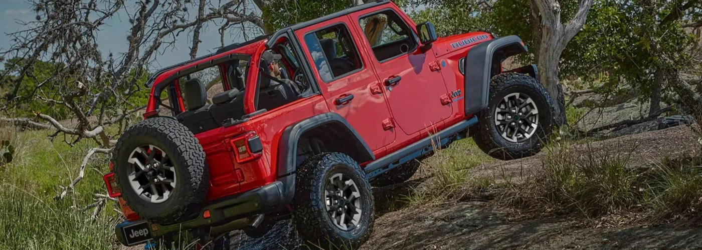 Red Jeep Wrangler 4-door off-road vehicle with open doors parked on rocky terrain surrounded by trees and brush.